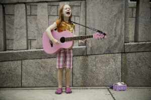 A young girl busking with a pink guitar on a street corner