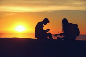 Two people playing the guitar silhouetted by a sunset in the background 