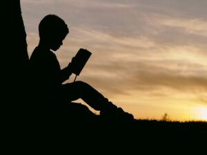 A young boy reading outside with a sunset inn the distance
