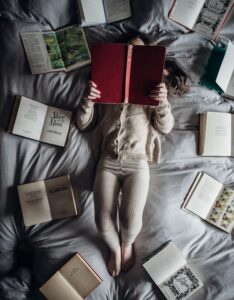 A child lying in bed reading surrounded by books